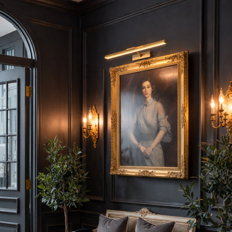 16" Wireless Brass Picture Light illuminating a classical portrait in a stately library; battery-powered art lighting with a CRI 90+ warm glow and brushed gold finish.